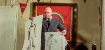 A still from the film Mis shows a bald man in a thick grey wool suit holding up two sketches of a giant rabbit type statue. He is standing in an office type room with a rainbow flag behind him.