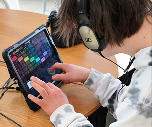 A boy sits with headphones on, playing on a tablet open on a music making app.