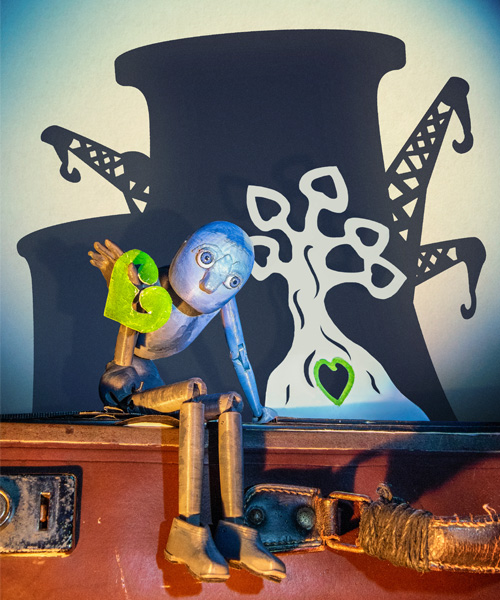 A wooden puppet with large eyes sits on top of an old suitcase. It is holding a green paper leaf in its left hand, shaped like a heart. Behind it are the silhouettes of two power plant towers and three cranes, over-laid with the illustration of a white tree.