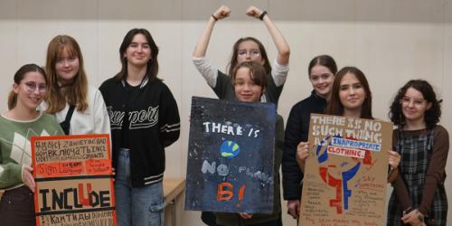 A group of young people hold protest signs against a plain wall