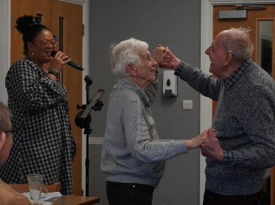 Two older people dance while a woman sings in a community hall